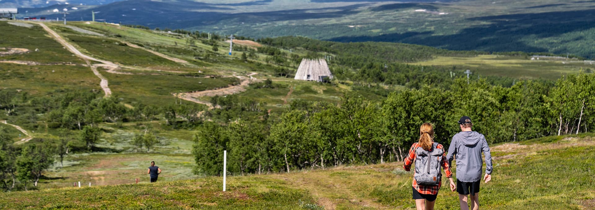 Två personer vandrar på en stig i fjällen, med gröna ängar och fjäll i bakgrunden. En lift och en arkitektonisk byggnad syns på avstånd.