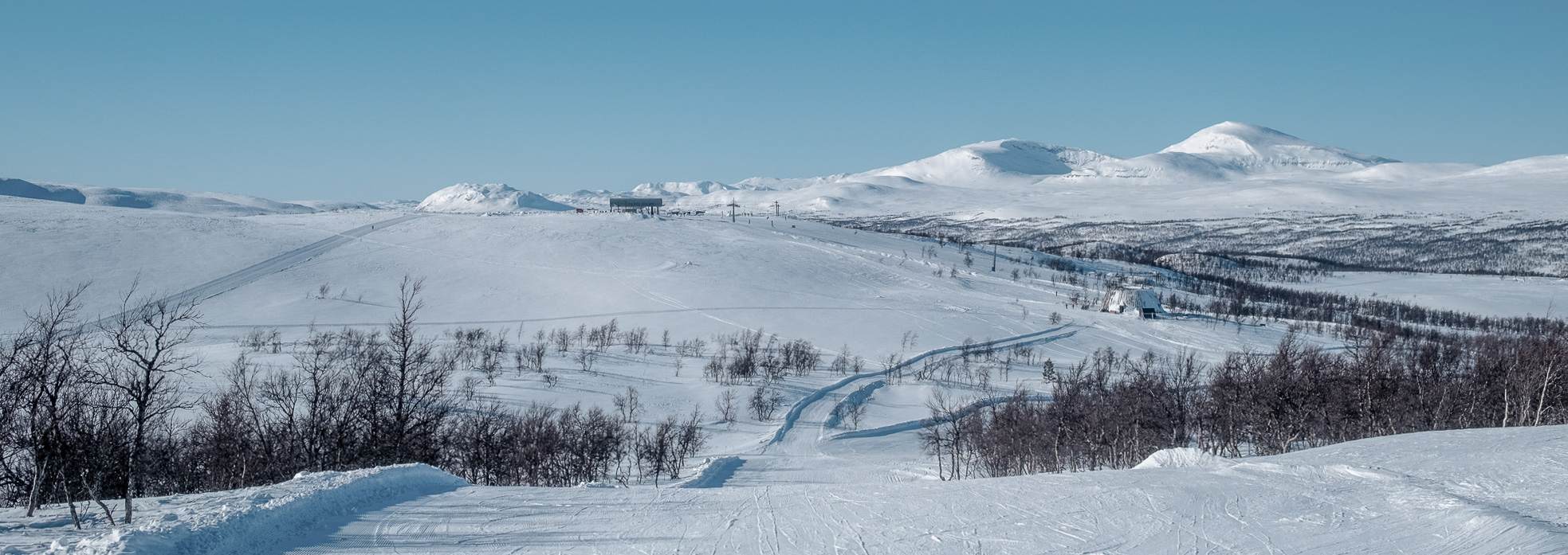 Två personer vandrar på en stig i fjällen, med gröna ängar och fjäll i bakgrunden. En lift och en arkitektonisk byggnad syns på avstånd.