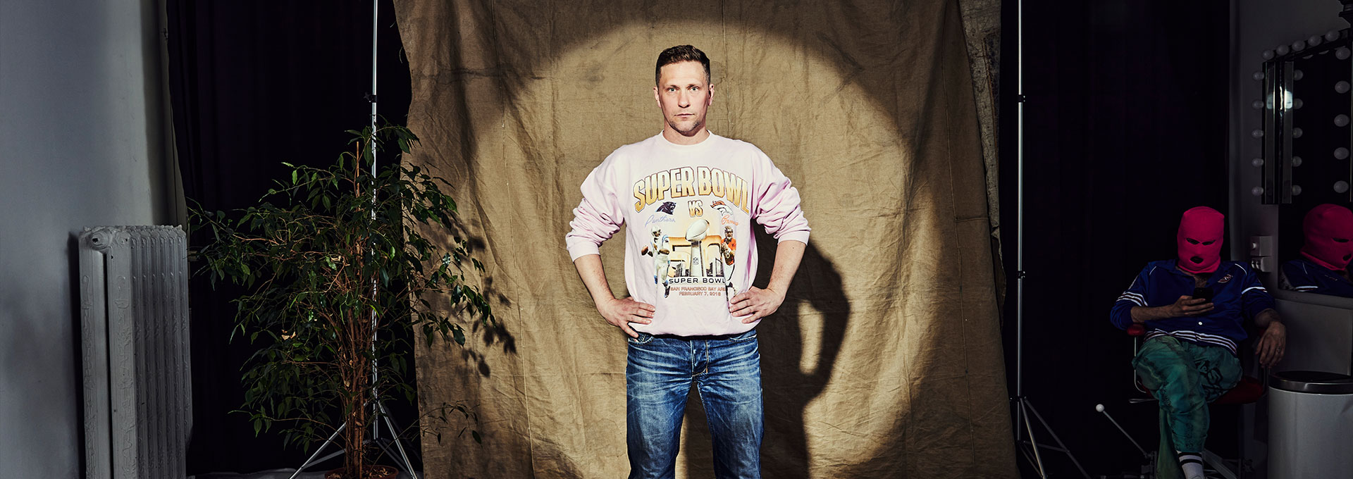 Komikern Magnus Betnér står i strålkastar ljuset med armarna på höften framför en tygbakgrund