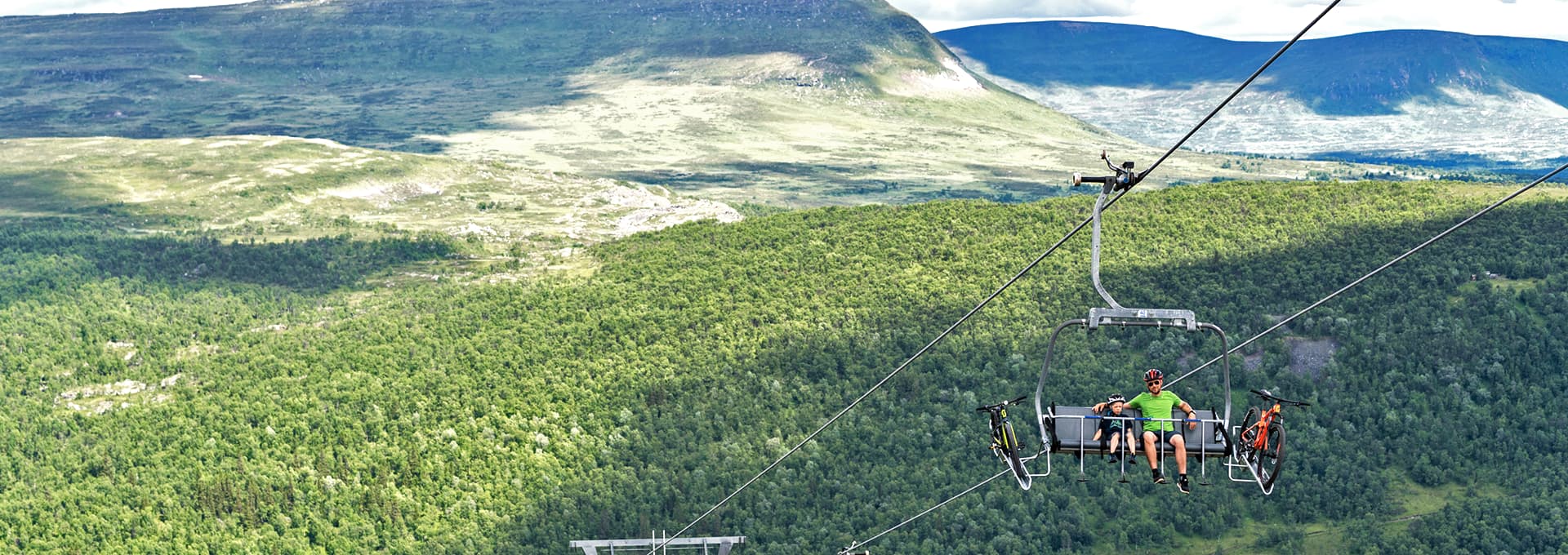Två personer vandrar på en stig i fjällen, med gröna ängar och fjäll i bakgrunden. En lift och en arkitektonisk byggnad syns på avstånd.